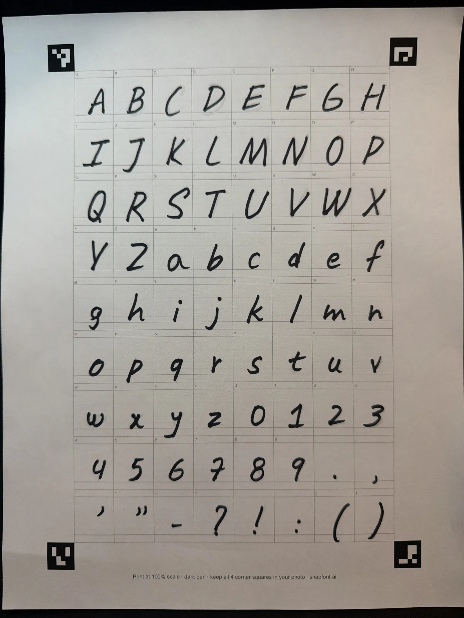 A photo of the SnapFont 72 box template filled in by hand with a black fine tip marker: capital letters A to Z, lowercase a to z, digits 0 to 9, and punctuation, one per box, with the four black corner squares visible on a dark table